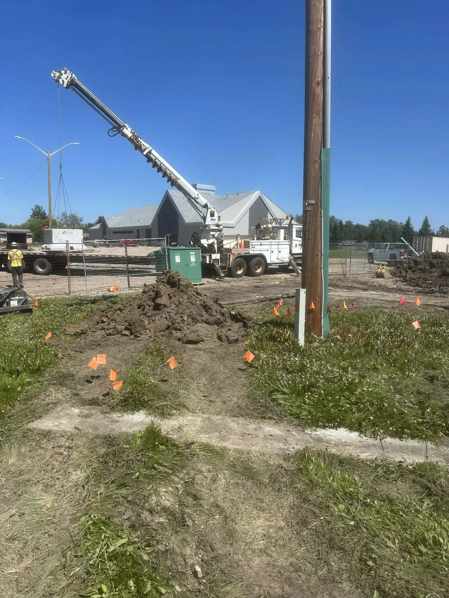 Construction site with a crane lifting a utility pole, workers, and dirt mounds, surrounded by orange markers and fences.