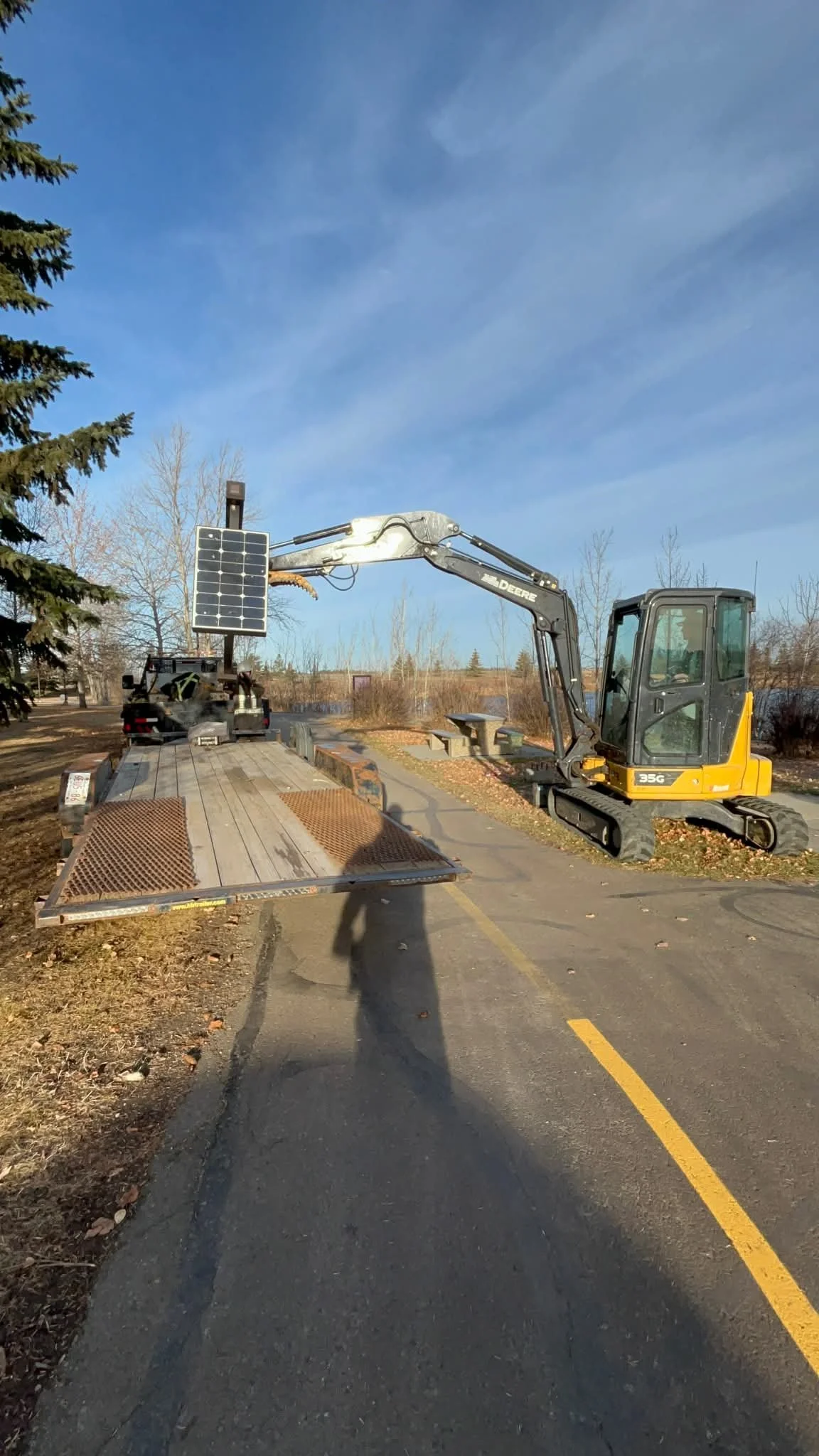 A small yellow and black excavator is lifting a solar panel onto a flatbed trailer in an outdoor parking lot on a sunny day with blue sky and some trees in the background.
