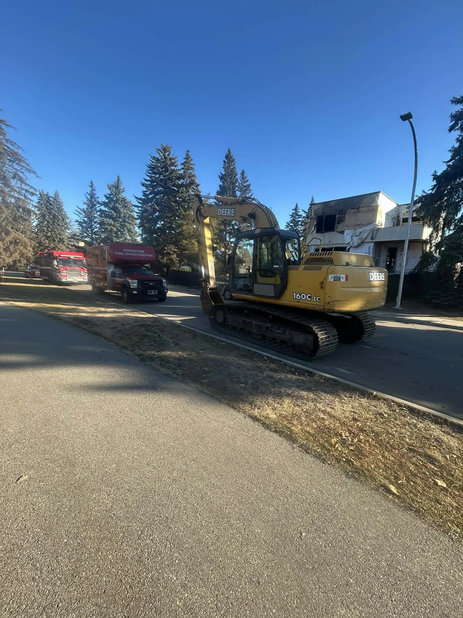 Construction excavator parked on the side of the street with emergency vehicles and a partially damaged building in the background.