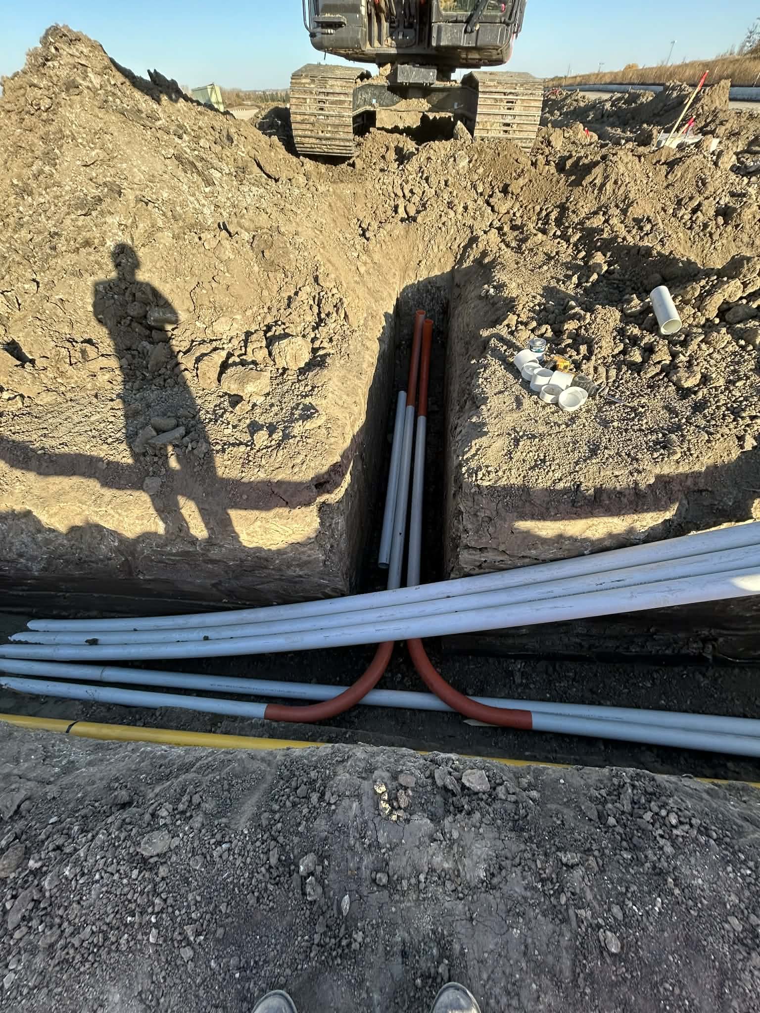 Construction site with exposed pipes and pipes laying in a deep trench, excavator in the background, and a person's shadow on the dirt.