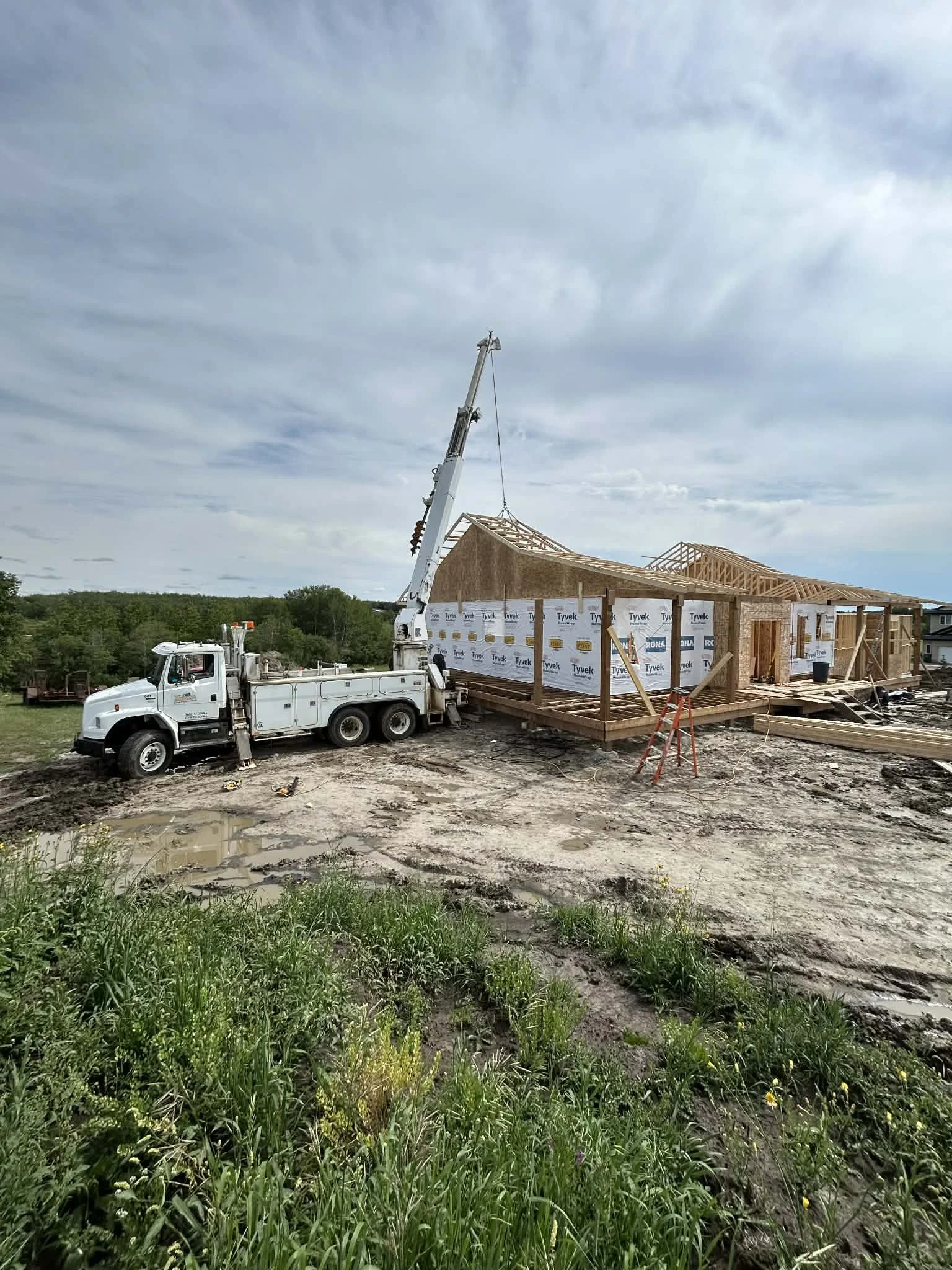 Construction site with a house being built, a crane lifting roofing materials, and a muddy ground with some green grass in the foreground.