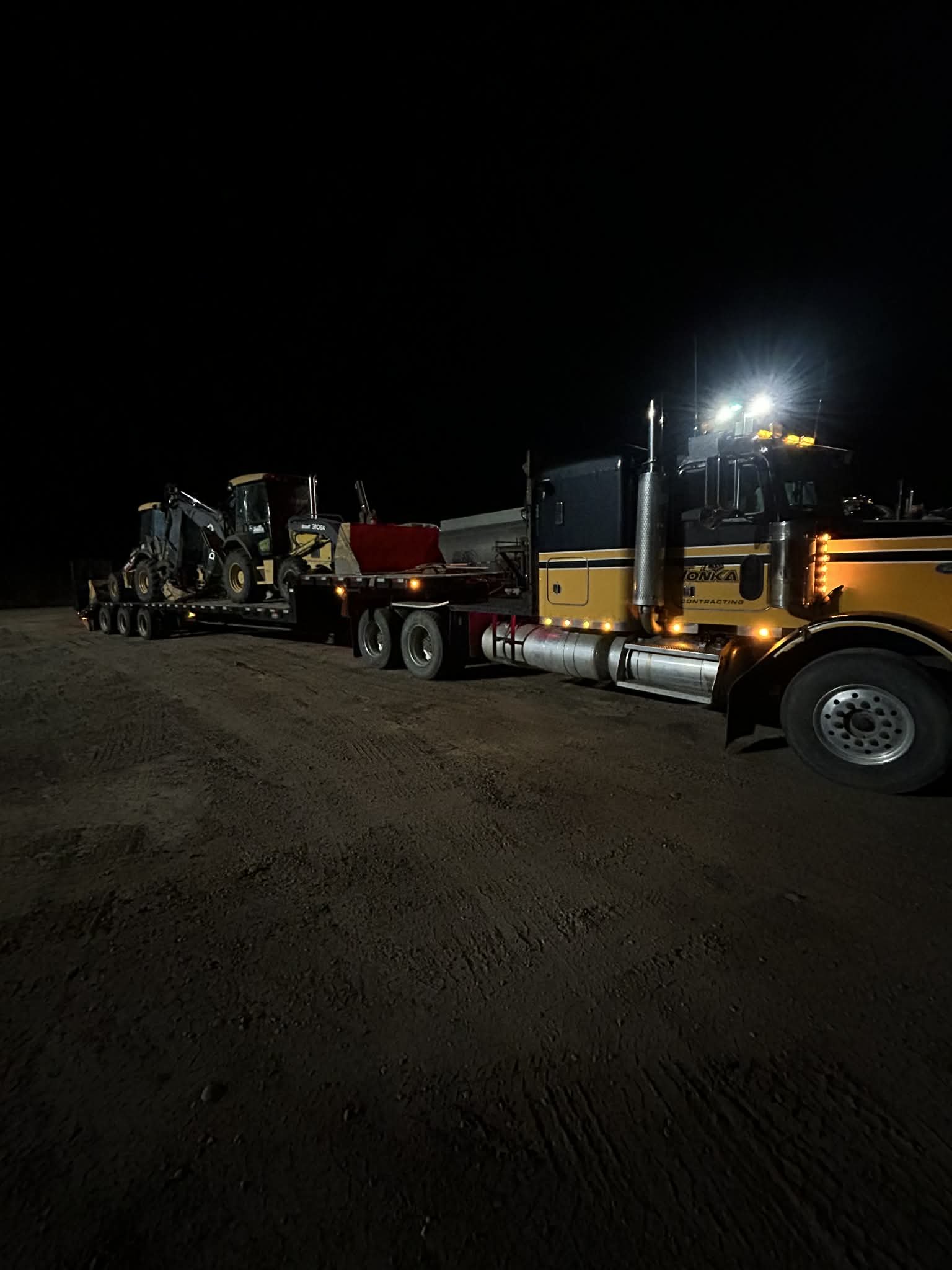 A large truck at night transporting construction equipment, including a skid steer loader and other machinery, on a dirt surface under bright lights.