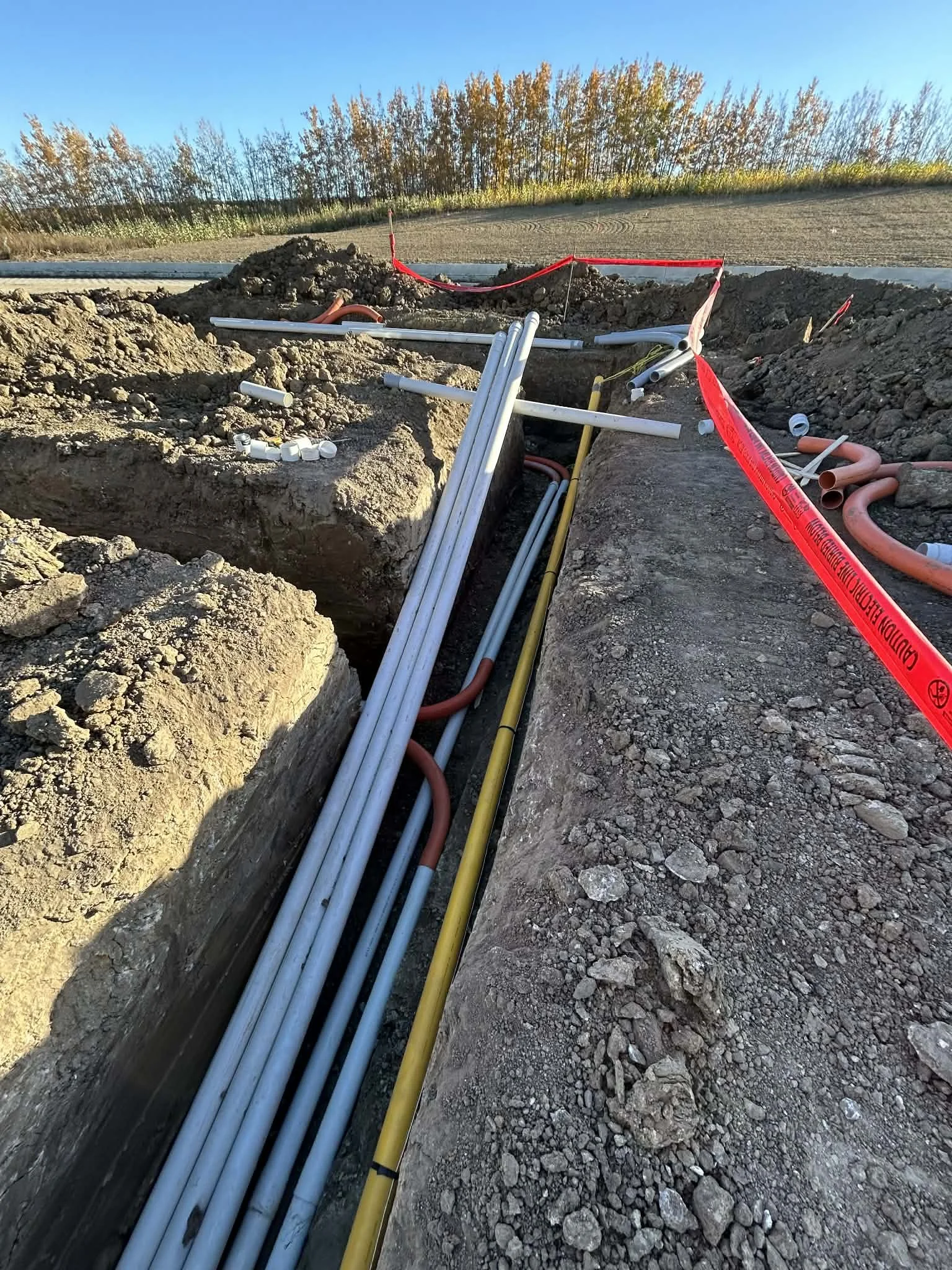 Underground construction site with pipes and cables in a trench, surrounded by red warning tape and open dirt, with a row of trees in the background.
