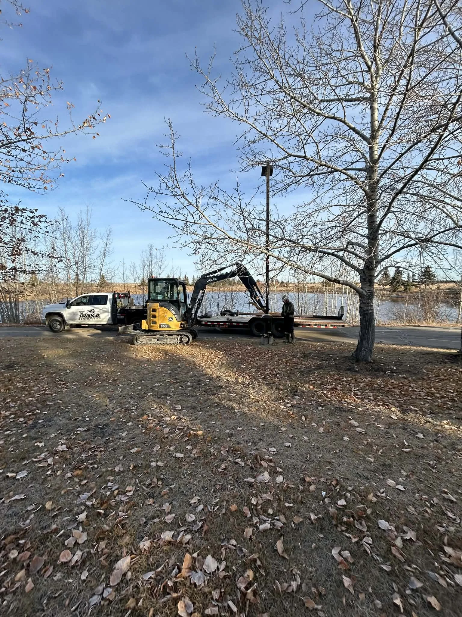 A small construction excavator on a trailer hitched to a pickup truck near a leafless tree, with a person standing next to them, by a body of water on a clear day.