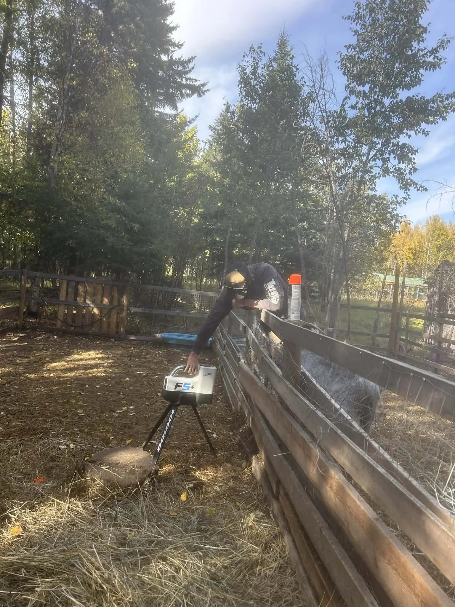 A person wearing a black jacket, jeans, and a helmet is working outdoors in a fenced area with trees and a blue sky, using a piece of equipment placed on a tripod.