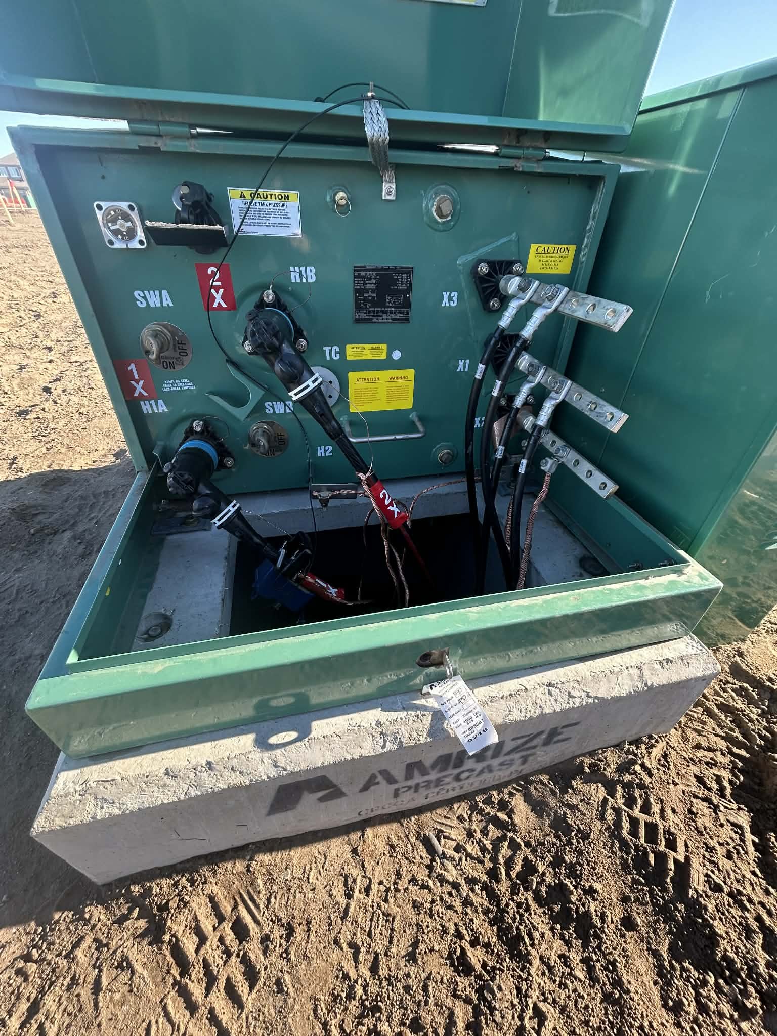 Open green electrical or control box with wiring and connectors inside, mounted on a concrete base on dirt ground, with labels and caution stickers on the inside panels.