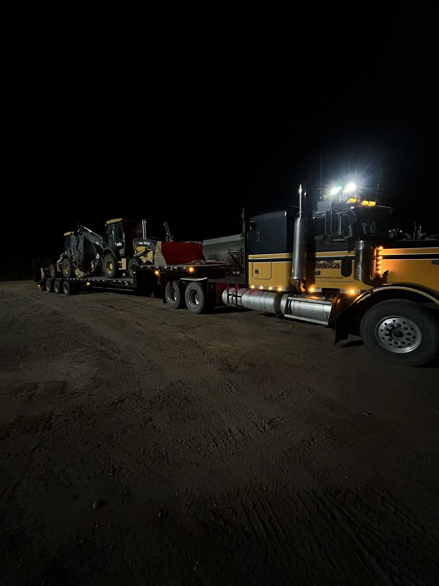 A large semi-truck carries construction equipment on a flatbed trailer at night, with bright lights illuminating the vehicle.