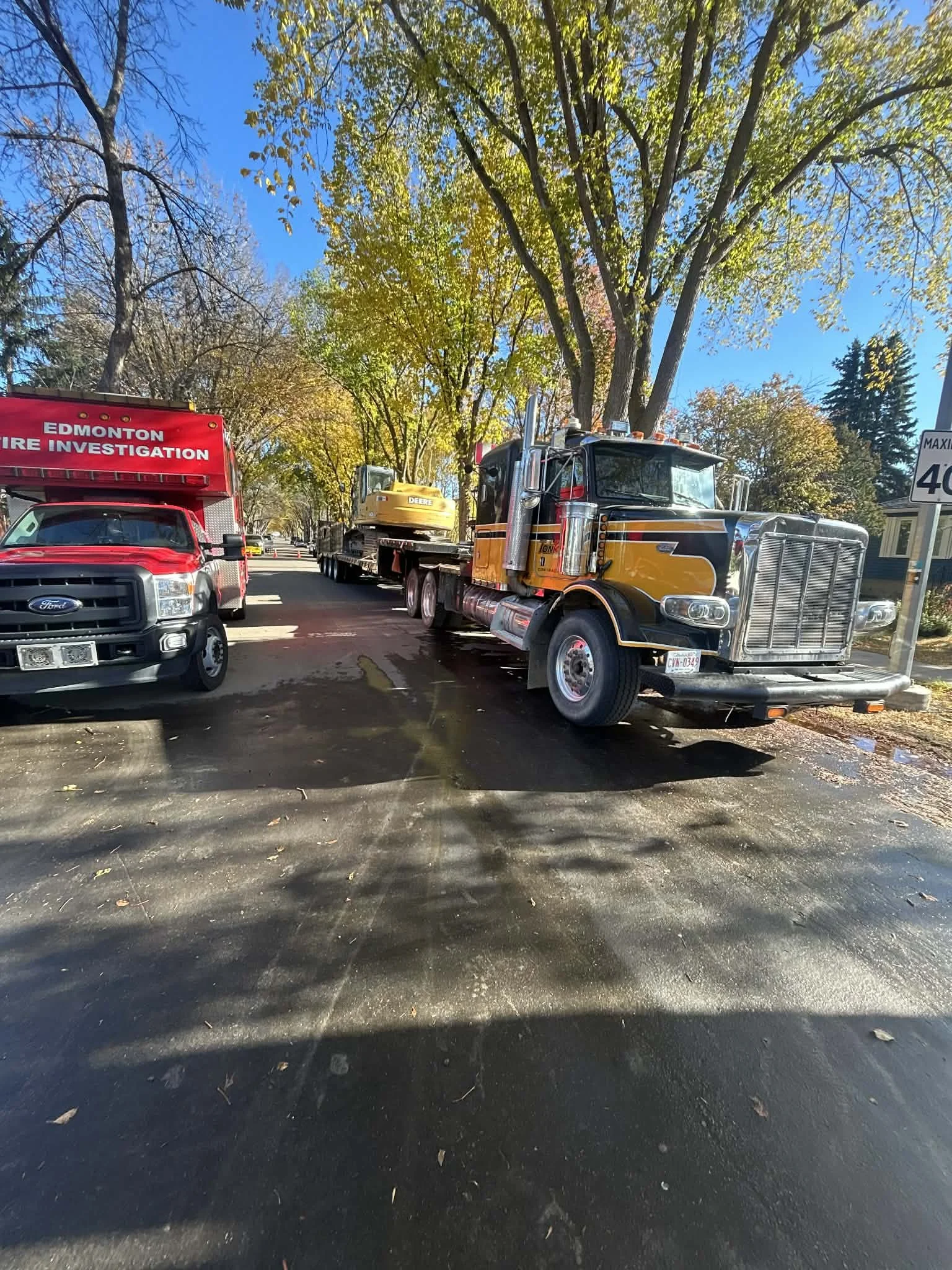 A street scene with a red Edmonton Fire Investigation truck and a yellow and black semi-truck carrying construction equipment parked on the side of the road. There are trees with green and yellow leaves overhead and a clear blue sky.