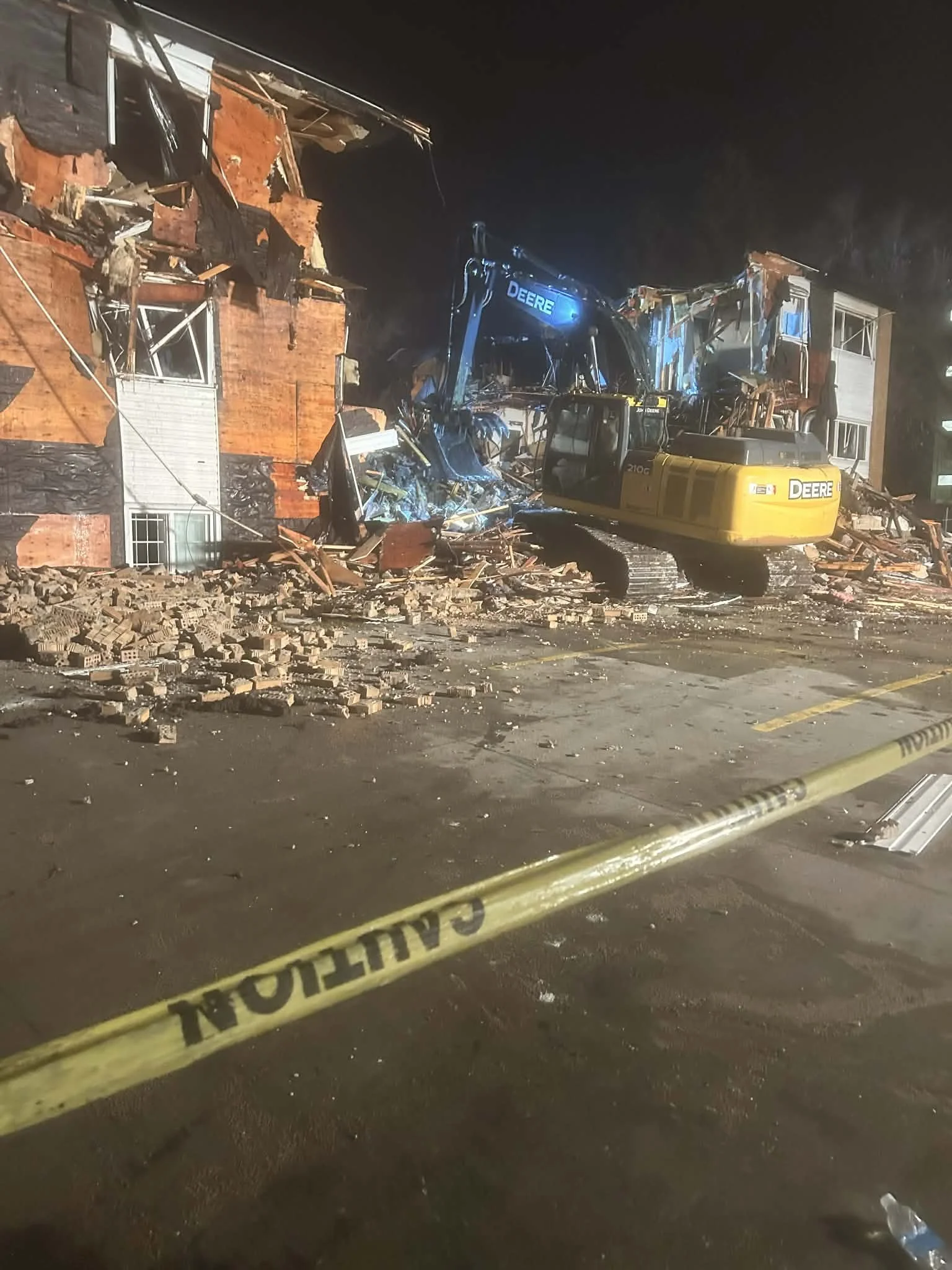 Construction vehicle demolishing a building with debris scattered on the ground, police caution tape in the foreground at night.