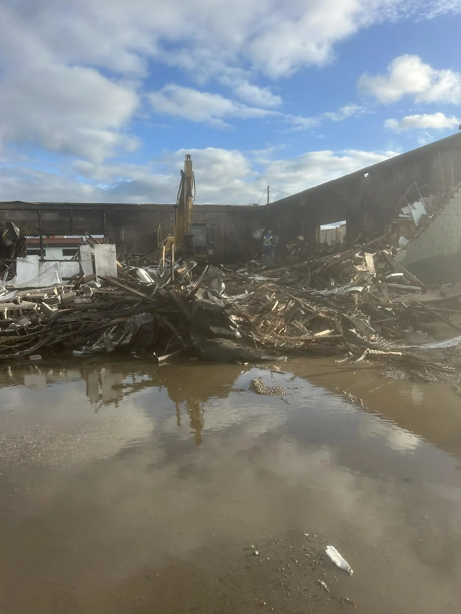 Remains of a building destroyed by fire with debris scattered, muddy water in the foreground, and a partially damaged wall with an opening, under a partly cloudy sky.