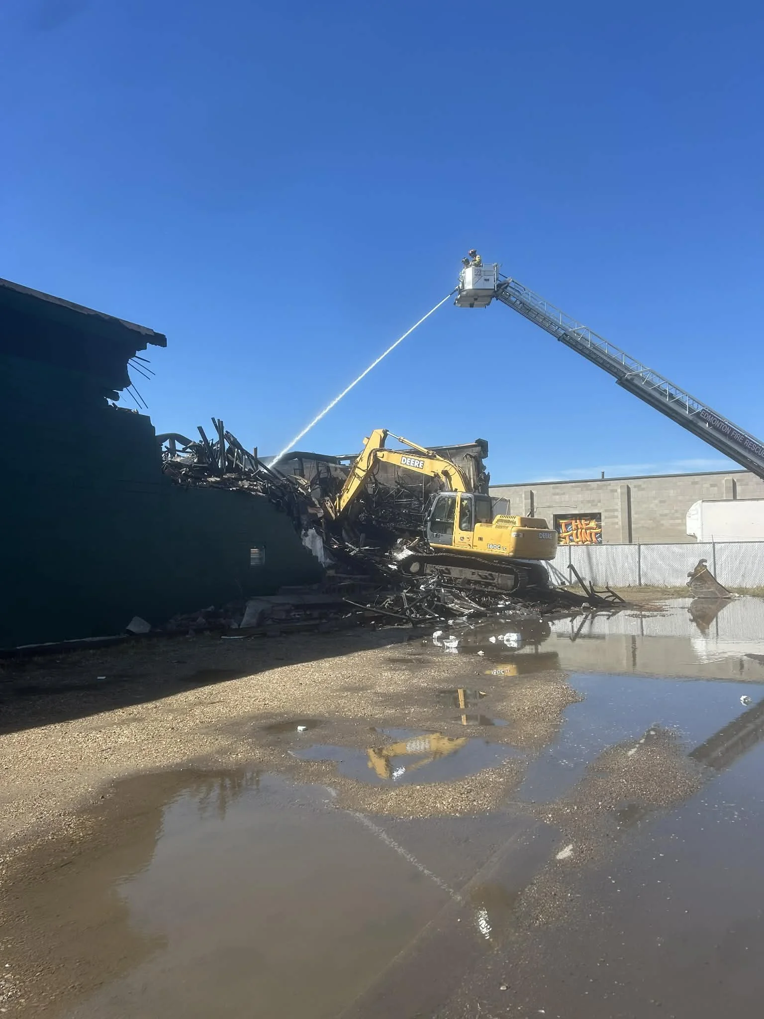 Firefighter on a ladder extending from a fire truck, spraying water on the wreckage of a damaged building, with a yellow excavator and a building in the background under a clear blue sky.