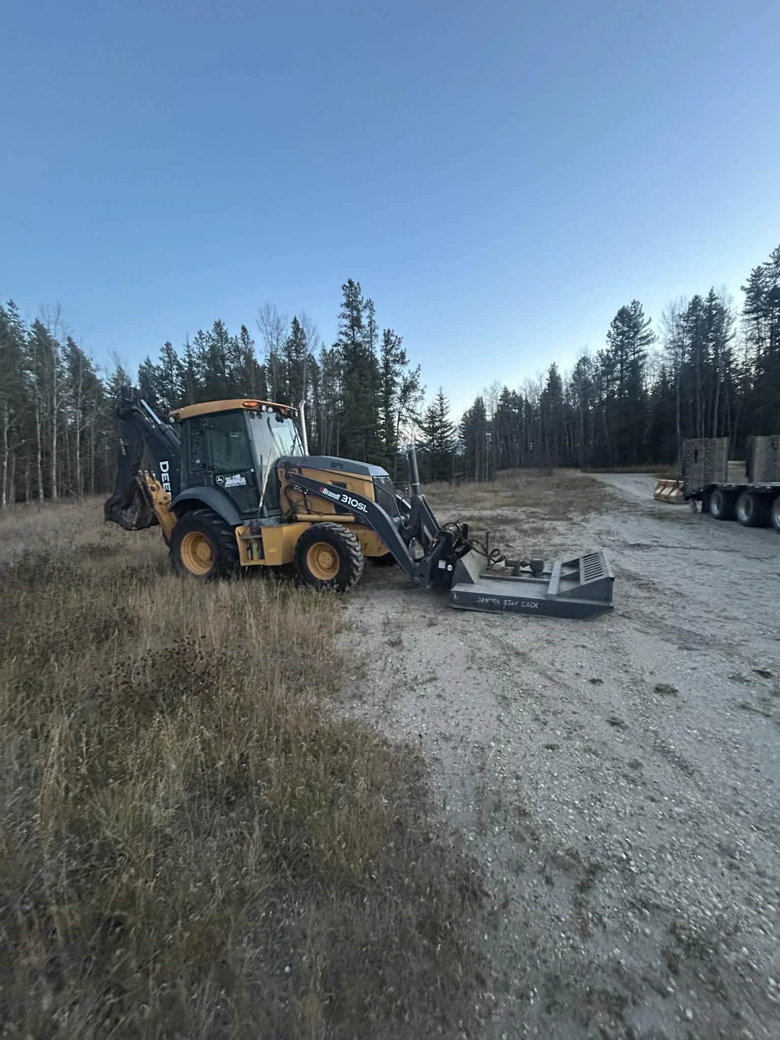 A John Deere tractor with a front loader attachment parked on a dirt road in a rural area, surrounded by trees under a clear blue sky.