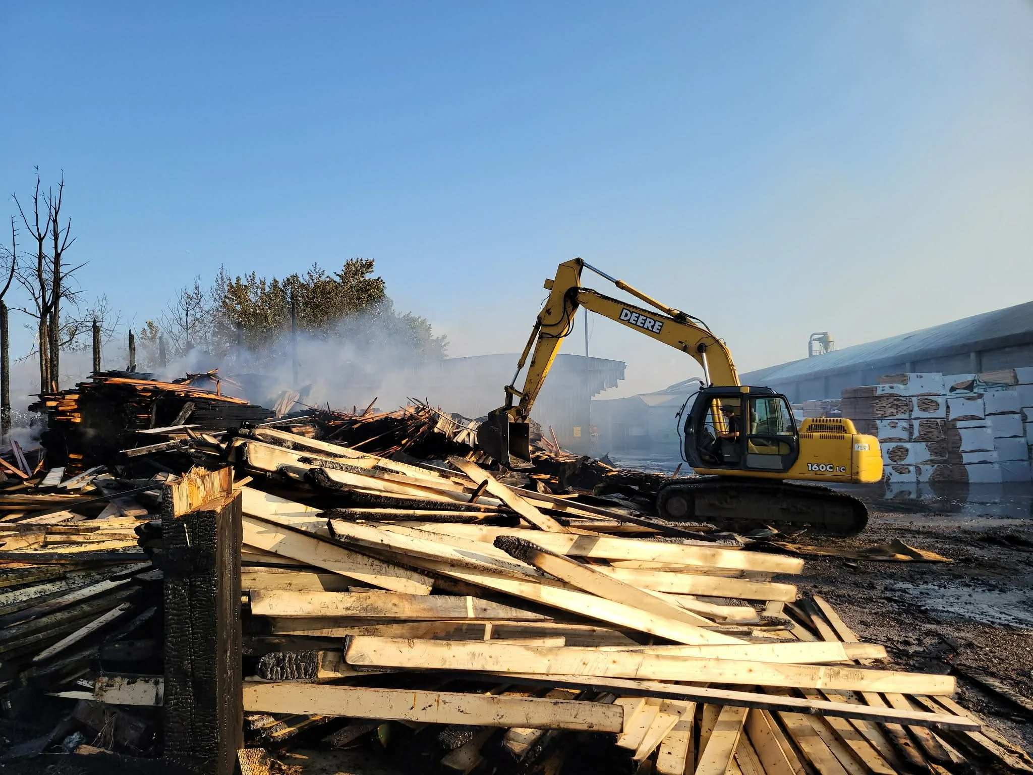 A yellow Deere excavator is demolishing a burned wooden structure, with smoke rising in the background and a clear blue sky.