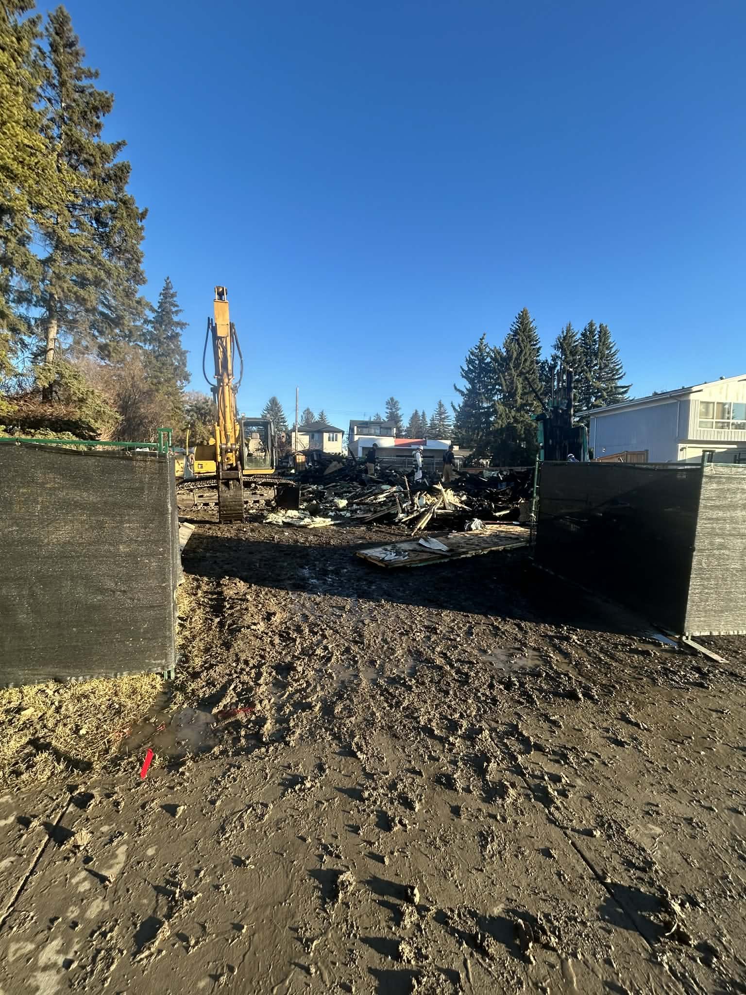 Construction site with an excavator and debris, surrounded by black fencing, with trees and residential buildings in the background under a clear blue sky.