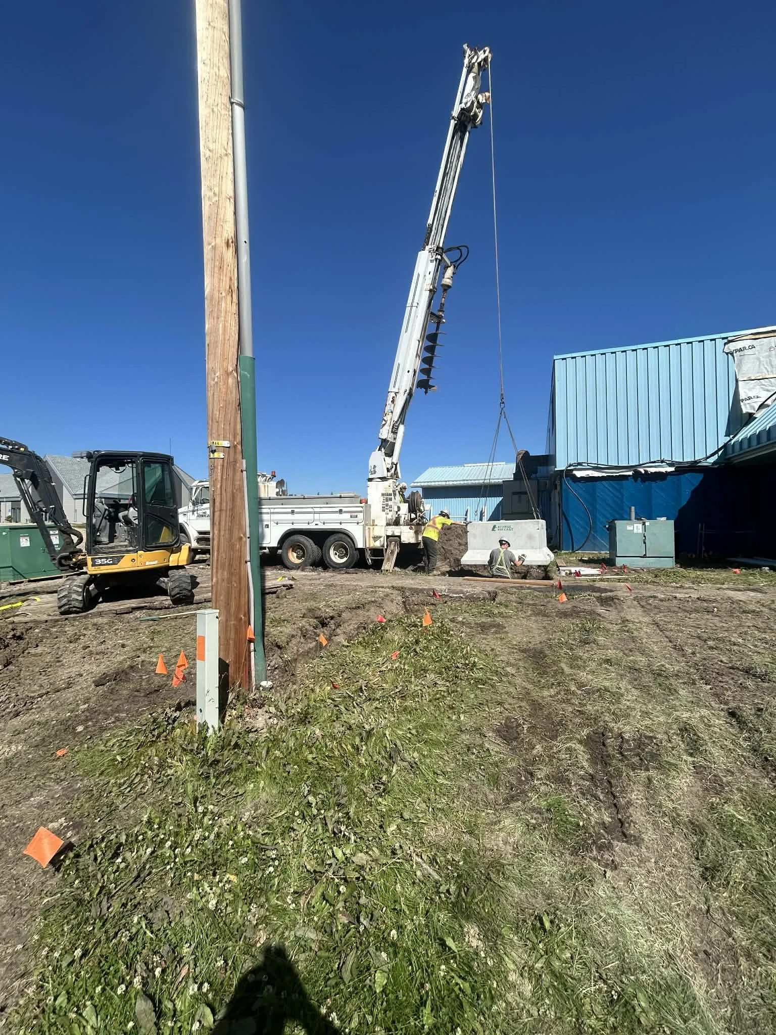 Construction site with a truck-mounted crane lifting a concrete barrier while workers operate nearby. There is a utility pole and a small excavator on the muddy ground.