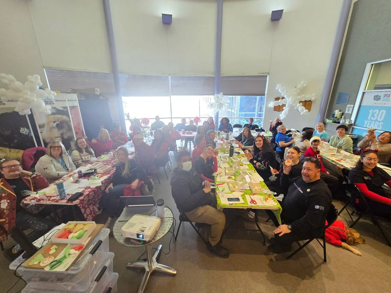Group of people gathered around decorated tables for a celebration in a bright room, with some wearing holiday attire and decorations.