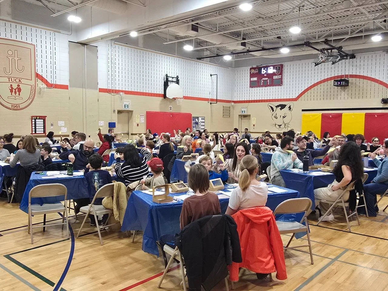 People sitting at tables with blue tablecloths in a gymnasium, participating in a group event or meal.
