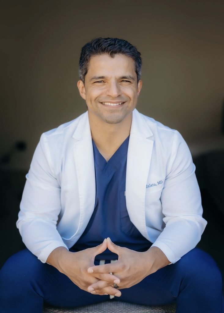 Andres Zuleta, MD, physician in a white lab coat embroidered Zuleta, MD, smiling with hands clasped