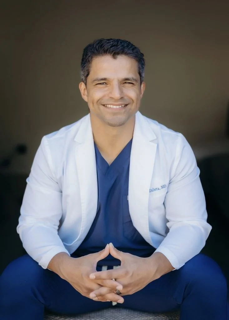Andres Zuleta, MD, physician in a white lab coat embroidered Zuleta, MD, smiling with hands clasped