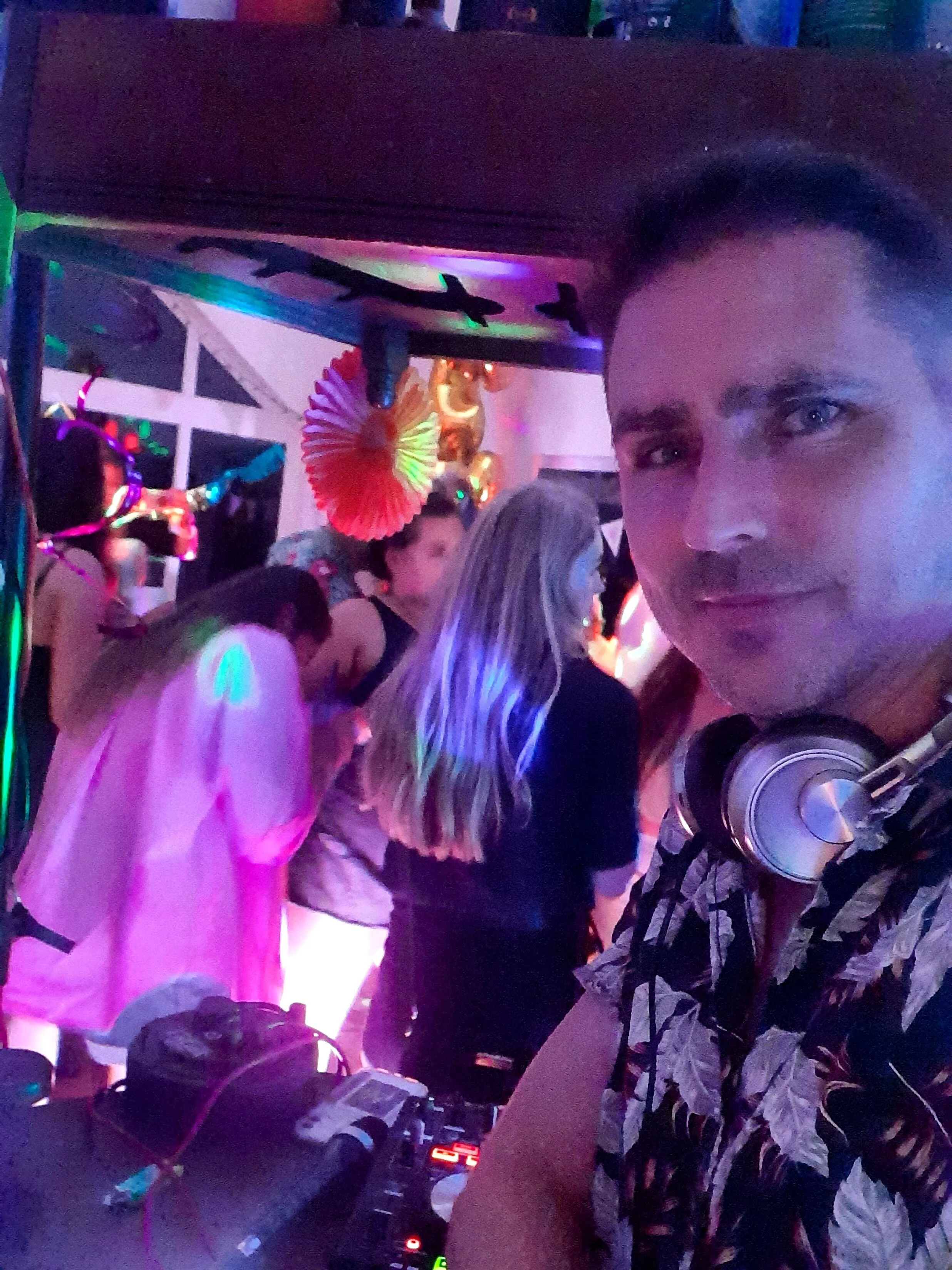 A DJ with headphones and equipment at a lively party with colorful lights and decorations.