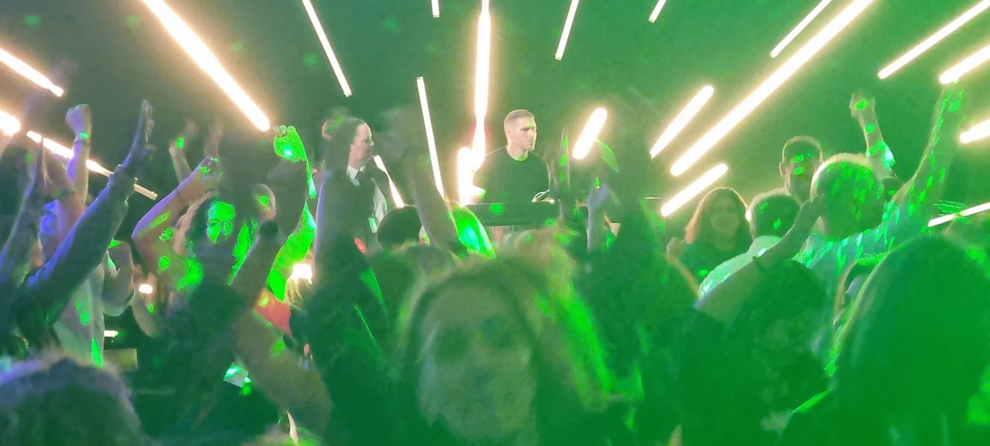 A vibrant nightclub scene with a DJ performing on stage. The crowd of people is dancing with raised arms, surrounded by green laser lights and bright linear light effects.