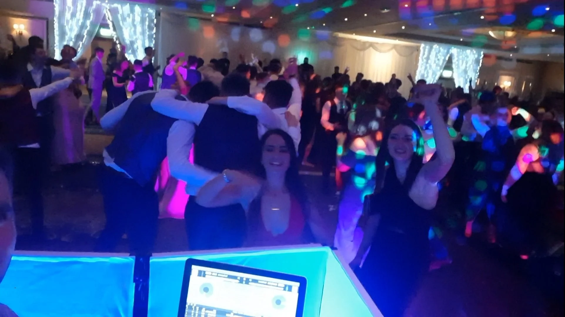 Crowded dance floor with colorful lights, people dancing, and a DJ booth visible in the foreground.
