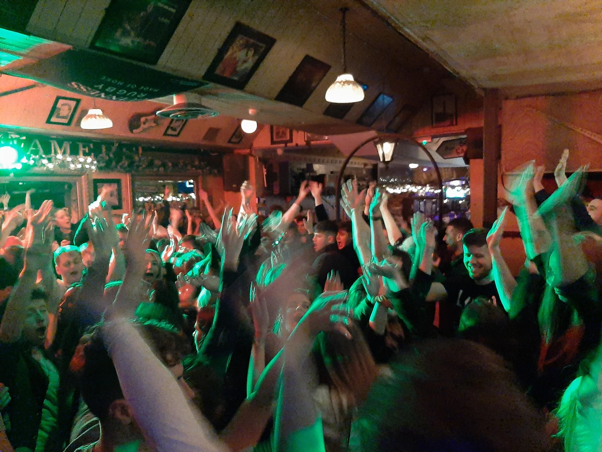 Crowd of people cheering and raising hands in a lively indoor event or party, with warm lighting and decorations in the background.