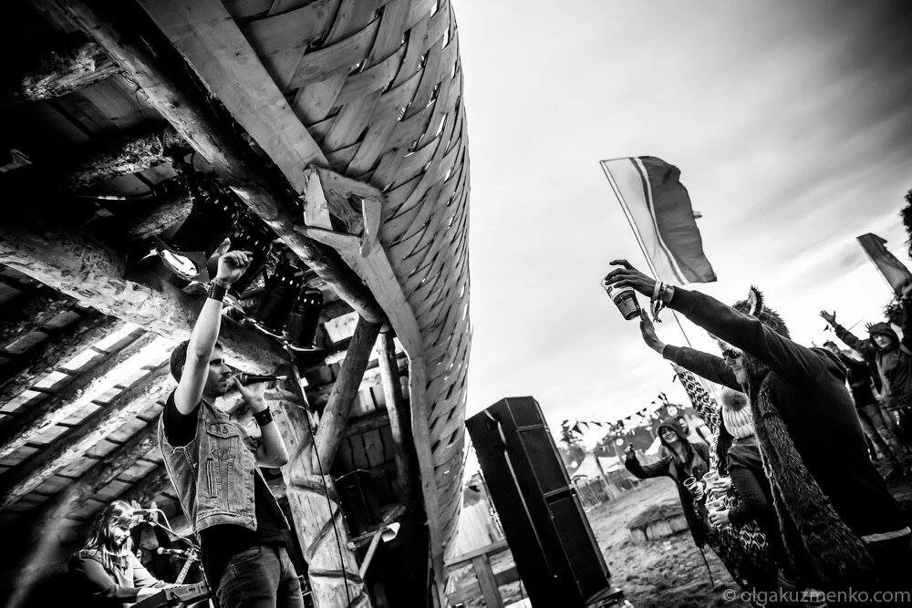 Black and white photo of a lively outdoor concert scene with enthusiastic audience, a performer under a wooden structure, and raised hands with flags.