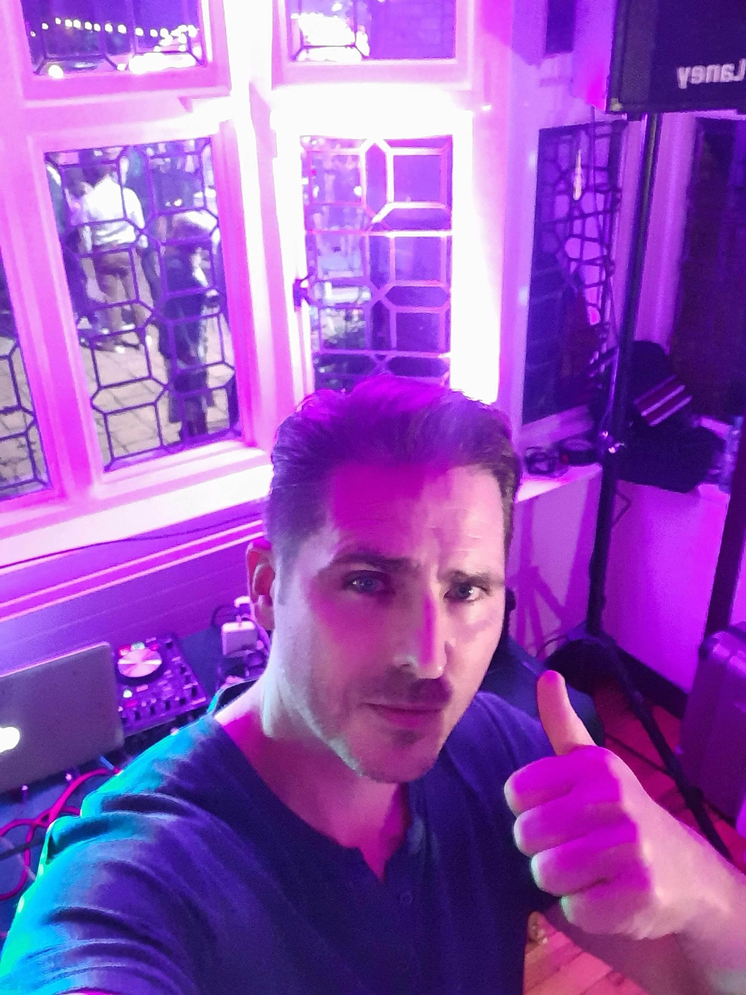 Person giving a thumbs up in a room with purple lighting, DJ equipment visible, and a window with people outside.