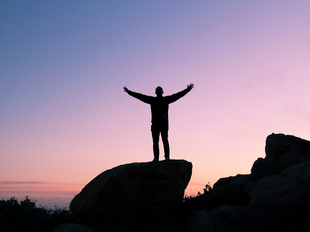 Person standing on a rock at sunrise with arms raised, representing a calm approach to January goal setting without starting over