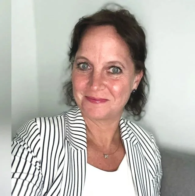 Eine Frau mit kurzen dunklen Haaren, trägt eine weiße Bluse mit schwarzen Streifen und eine Halskette, lächelt vor einem weißen Hintergrund.
