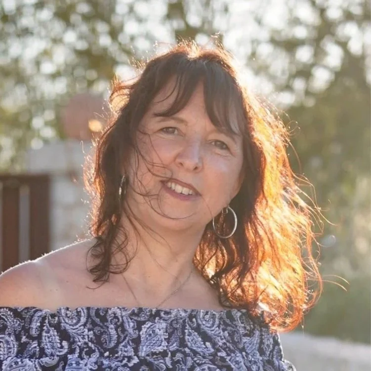 Frau mit dunklen, lockigen Haaren, trägt großen Ohrring und ein off-shoulder Kleid, im Sonnenlicht in einer natürlichen Umgebung.
