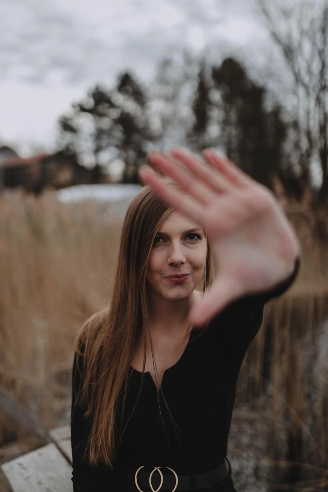 Junge Frau mit braunen Haaren in einer schwarzen top, die mit der Hand nach vorne greift, im Freien mit Bäumen im Hintergrund, an einem bewölkten Tag.