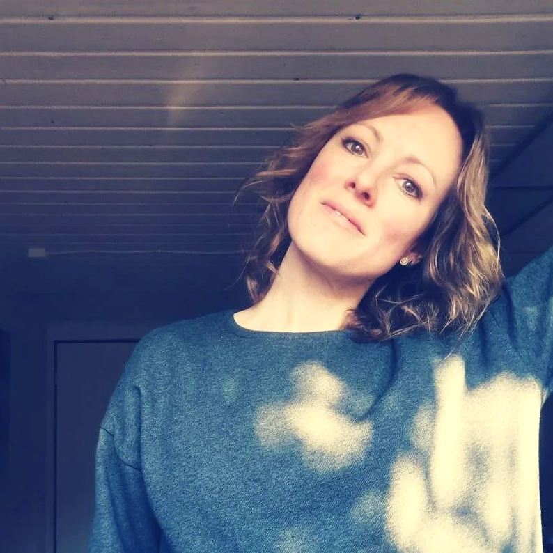 Eine Frau mit braunen, lockigen Haaren trägt einen grauen Pullover, steht vor einer Holzdecke und einem Türrahmen.