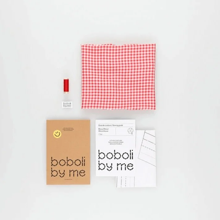 Conjunto de materiales para costura: una bolsa de tela con diseño de cuadros rojos y blancos, hilo color rojo, guía de costura, una tarjeta con el texto 'boboli by me', y un cuaderno o libro en color beige con una carita feliz en la esquina superior izquierda.