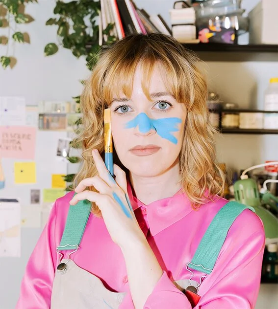 Una mujer con cabello rubio y ojos azules, con pintura azul en la cara, sosteniendo un pincel y luciendo ropa rosa y un overol. Está en un ambiente de oficina o estudio con plantas y notas en la pared atrás.