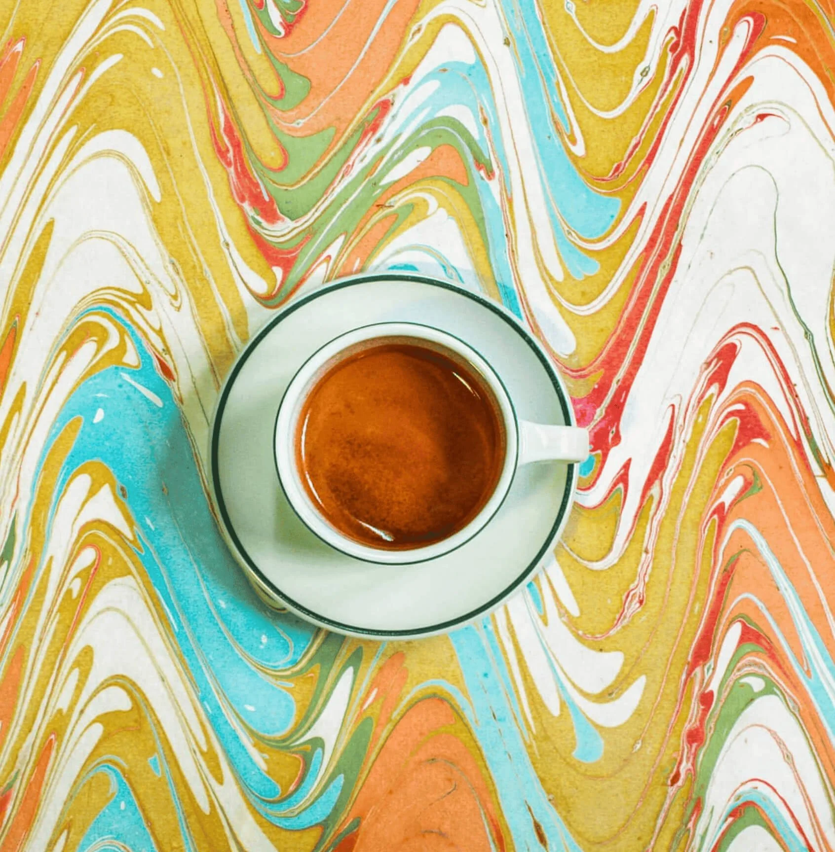 Taza de café sobre un plato en un fondo de mosaico de colores vibrantes con patrones de remolinos en tonos amarillos, azules, verdes, naranjas y rojos.