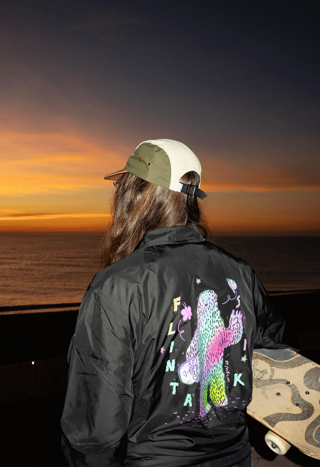 Persona vista de espaldas, con sombrero y chaqueta con diseño de dinosaurio y letras, en una escena junto a un mar al atardecer con cielo naranja y azul