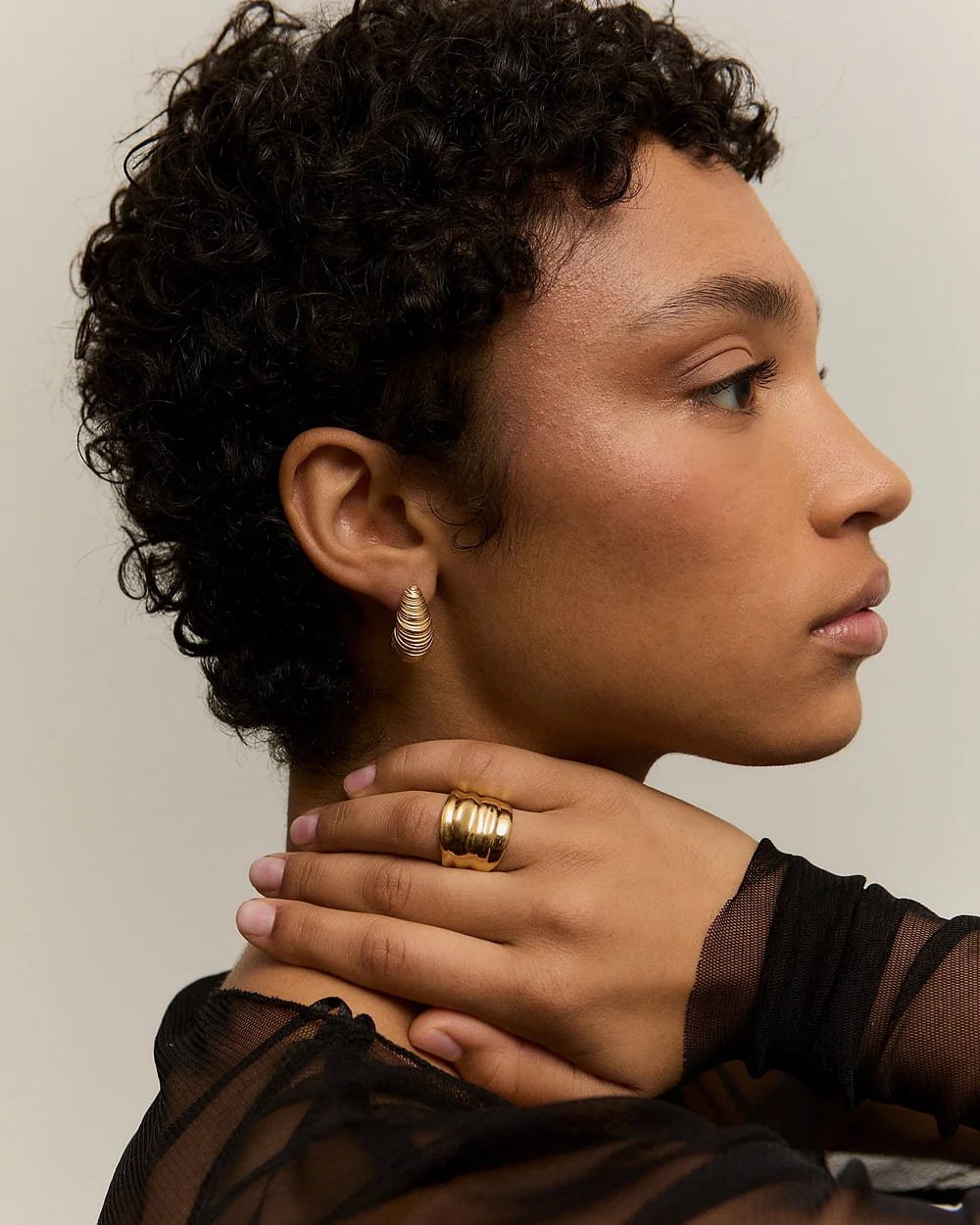 Retrato de perfil de una mujer con peinado rizado, usando anillo dorado en el dedo y pendientes de aro, vestida con blusa de malla negra, haciendo una pose elegante.