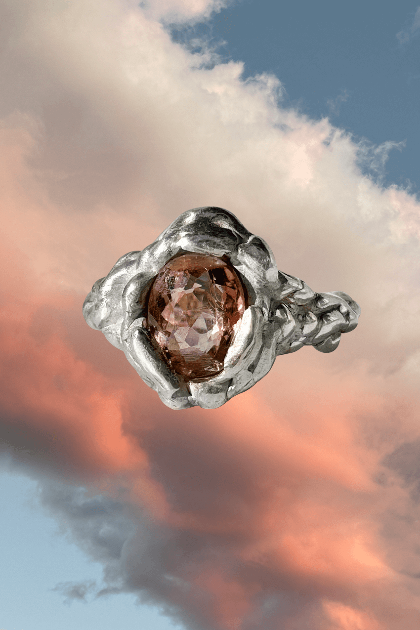 Anillo con piedra de color marrón en forma de óvalo, en un cielo con nubes y tonos rosados y azules.