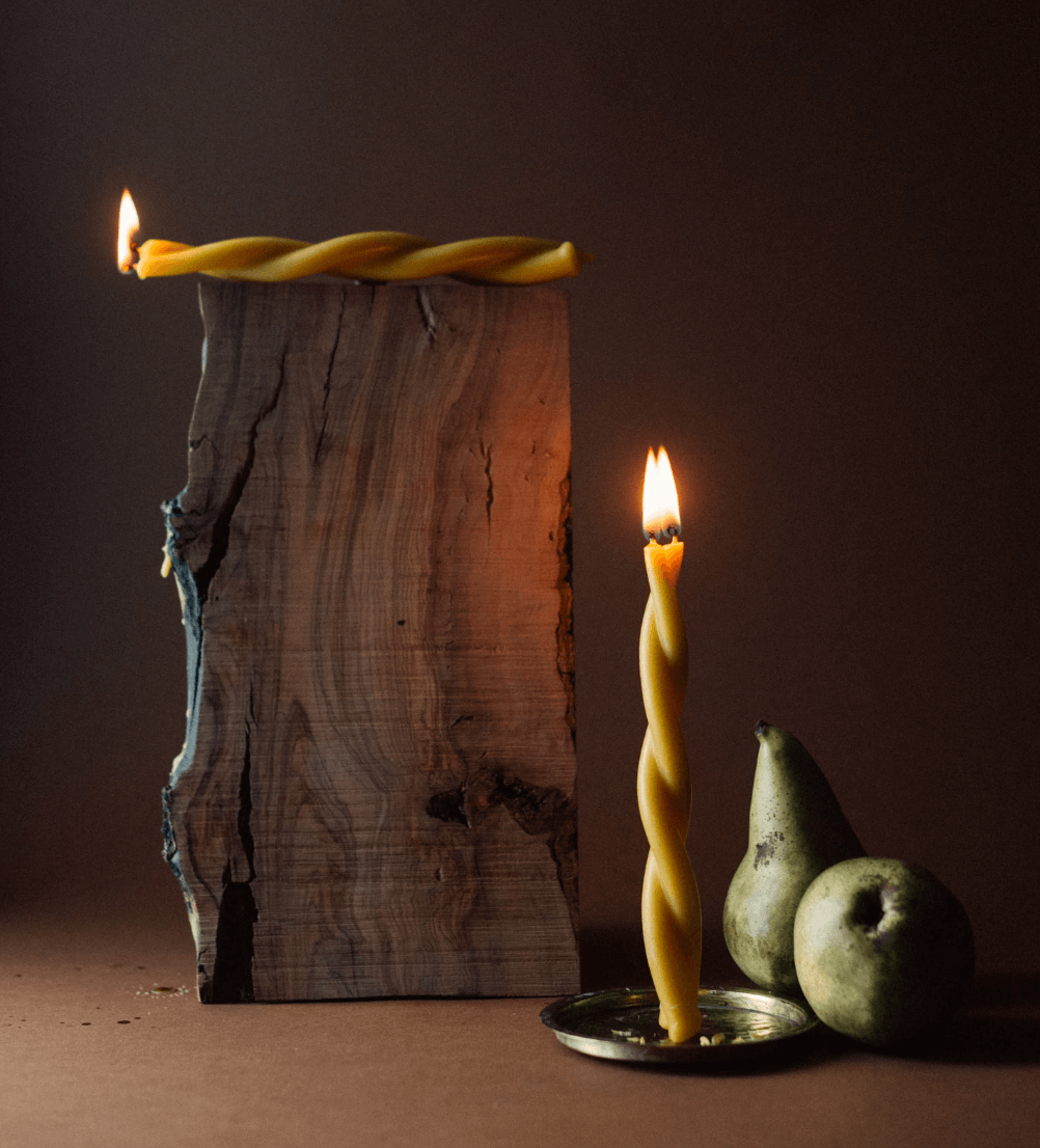 Vela torcida encendida, otra vela torcida también encendida, dos peras y un bloque de madera en un fondo oscuro.