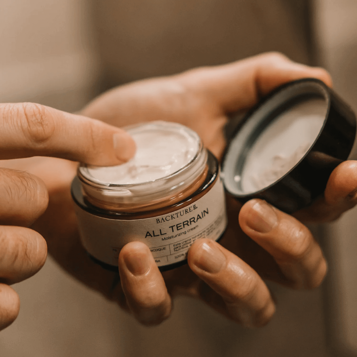 Manos abren un frasco de crema hidratante de la marca BACKTURE, llamado ALL TERRAIN.