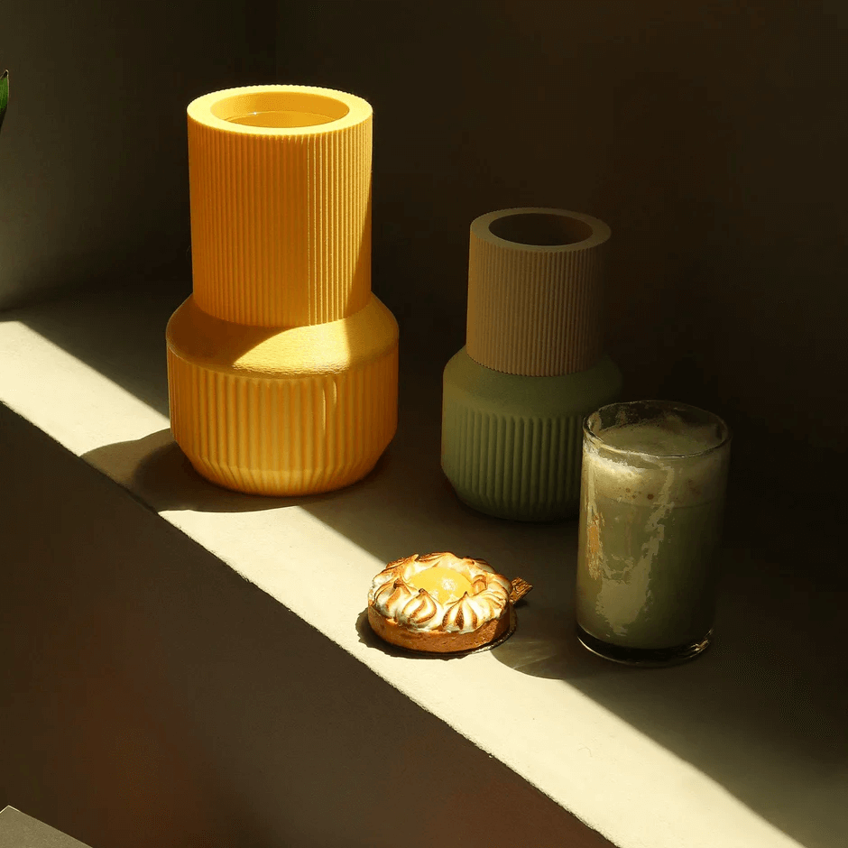 Cuatro objetos sobre una repisa: dos jarrones grandes, uno amarillo y uno verde, un pequeño pastel de crema y un vaso con leche