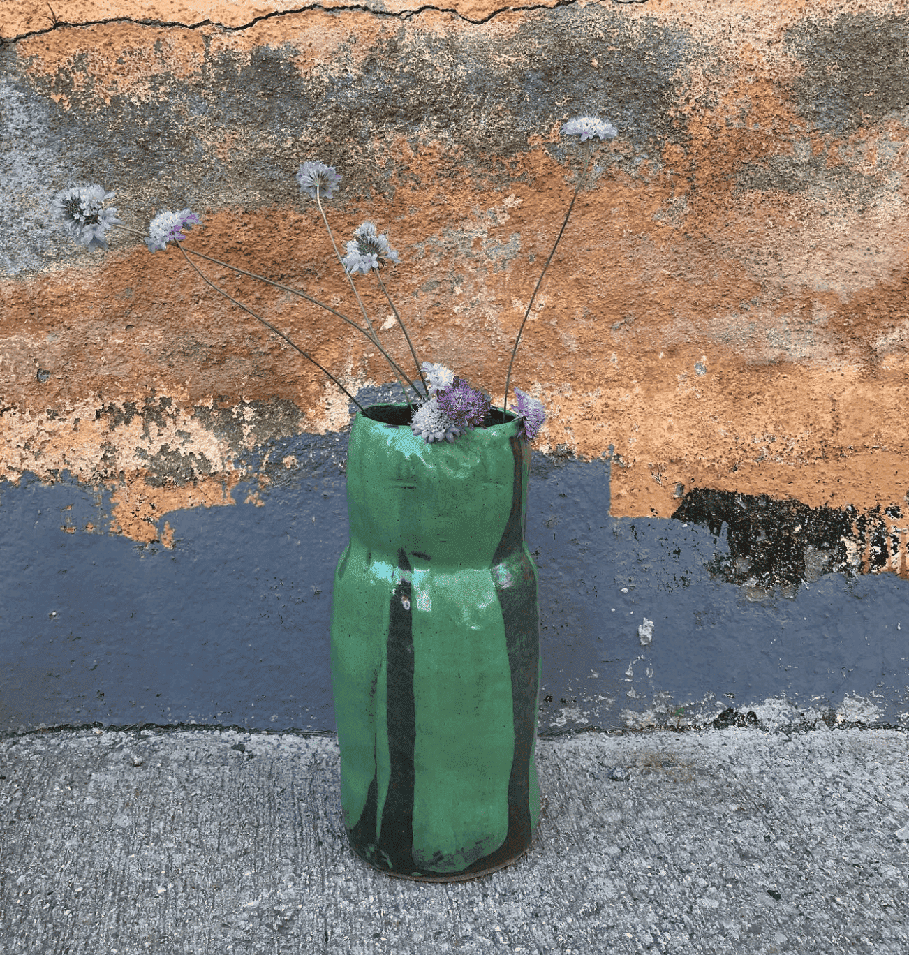 Vaso de cerámica verde con flores secas de color morado y gris, en un fondo de pared pintada en tonos anaranjados y grisáceos, y sobre un suelo de concreto.