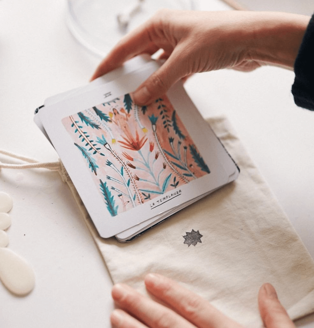 Persona sosteniendo una carta con ilustración floral y un poema, sobre una libreta de tela. Cerca hay unas ilustraciones blancas en la mesa.