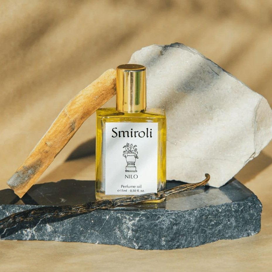 Frasco de perfume con etiqueta que dice 'Smirolli Nilo Perfume oil', rodeado de piedras y varillas decorativas en un fondo beige.
