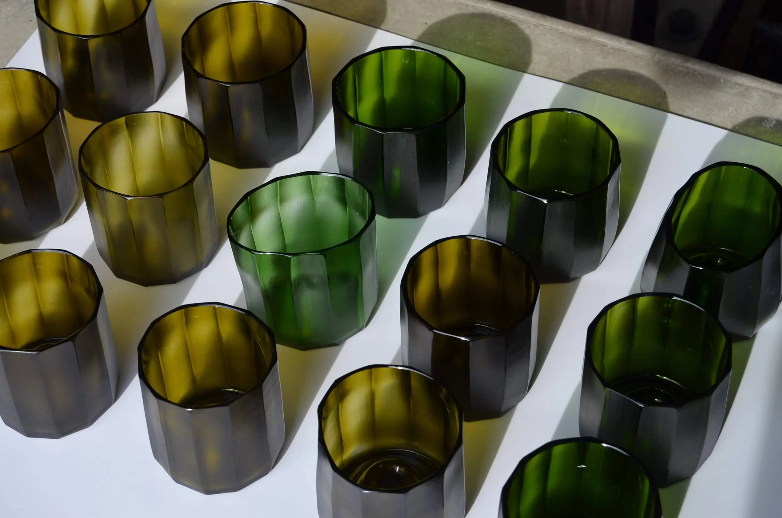 Vasos de cristal en colores verde y ámbar, colocados sobre una mesa blanca con sombras proyectadas.