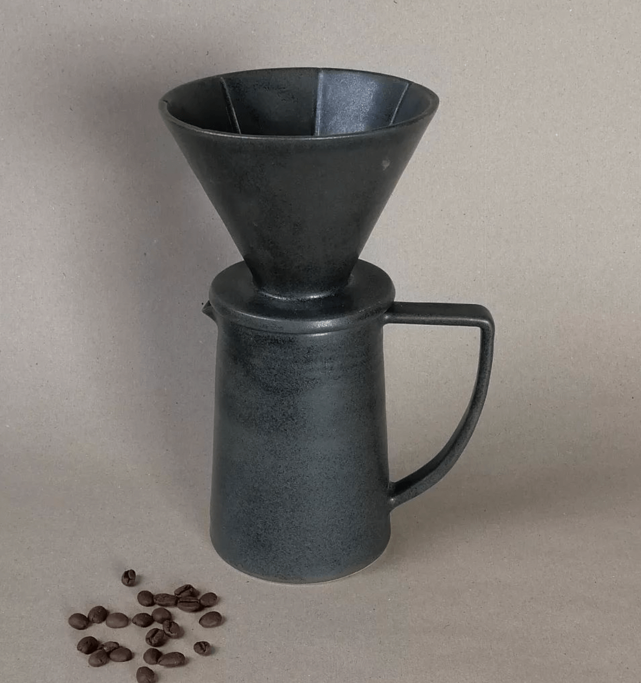 Prensa francesa, molinillo de café, con algunos granos de café a su lado, todo sobre un fondo gris claro.
