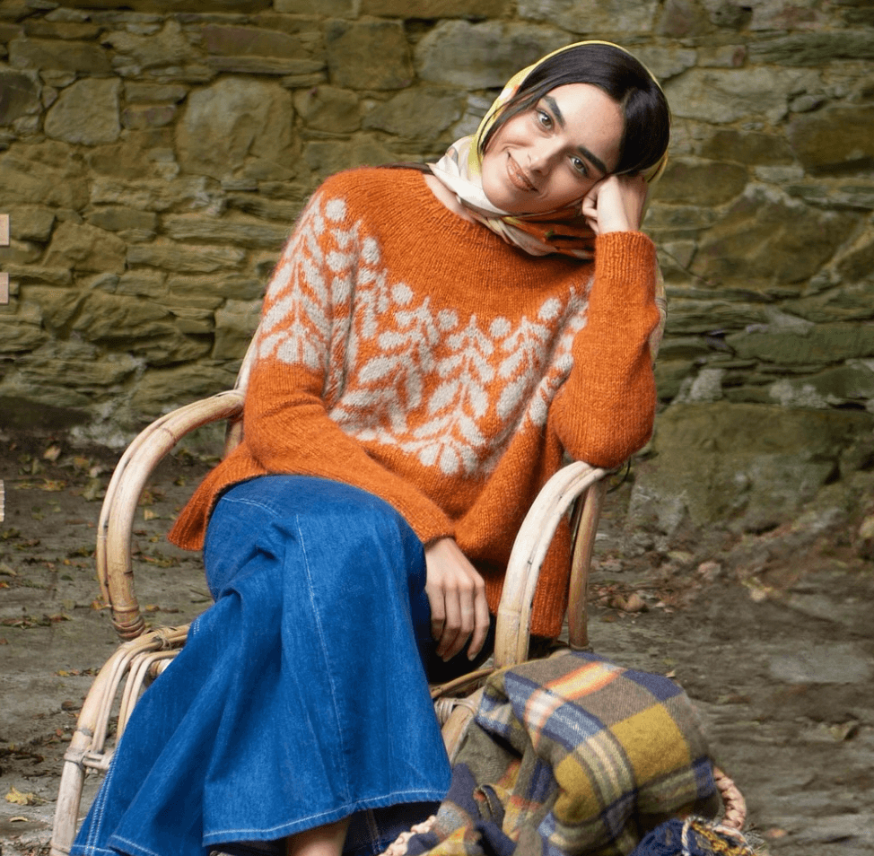 Mujer sentada en una silla de mimbre, con una bufanda de cuadros en las piernas, usando suéter naranja con diseño en color beige y jeans azul, sonriendo y apoyando la cabeza en su mano, con un fondo de muro de piedra.