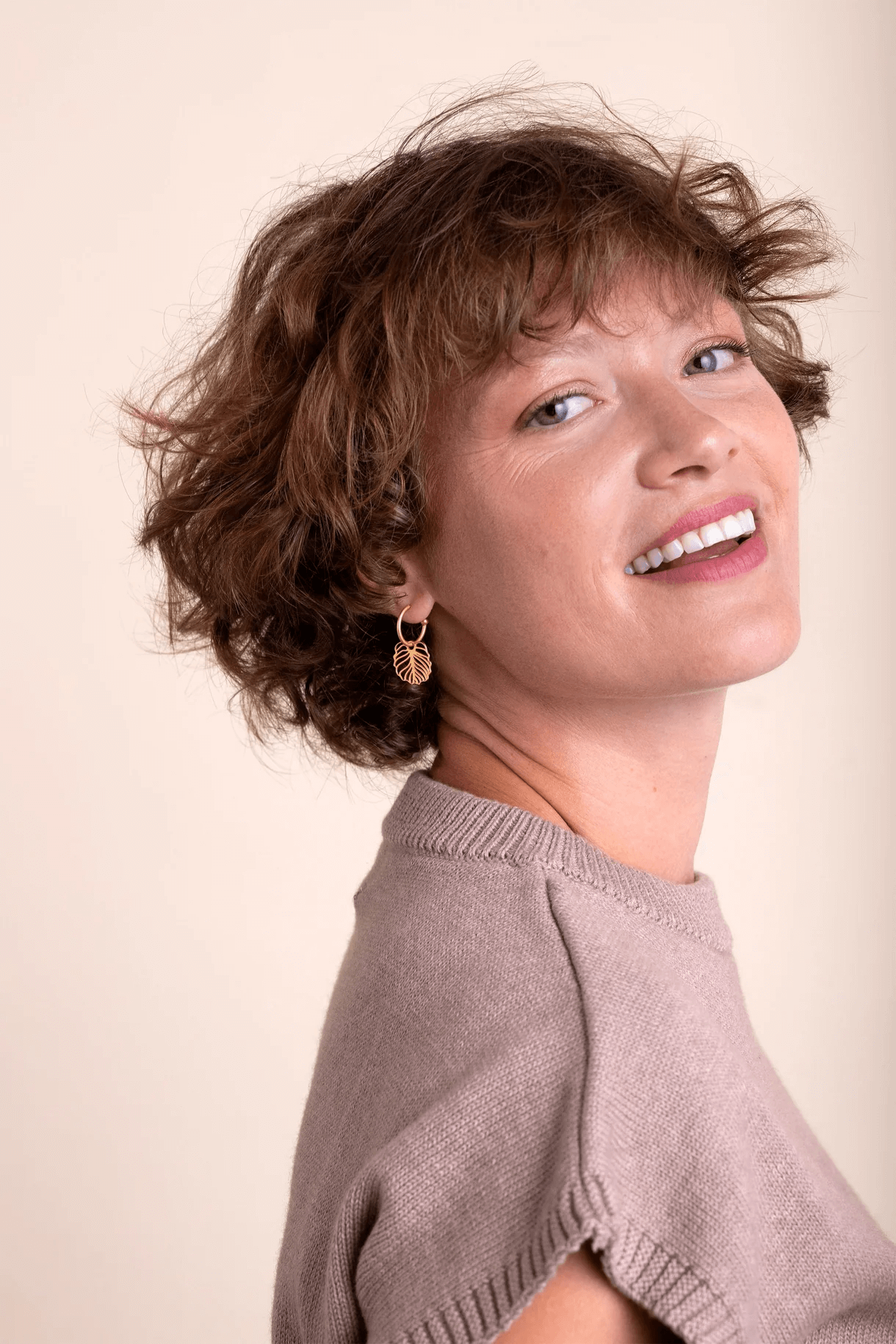 Retrato de una mujer con cabello rizado, piel clara, vistiendo un suéter gris y arete en forma de hoja, sonriendo con boca abierta.